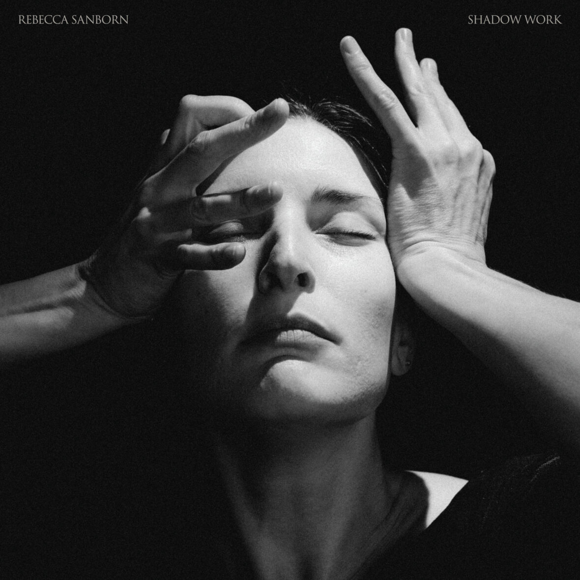A woman with an expressionless face in the shadows holding her hands to her face.