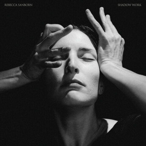 A woman with an expressionless face in the shadows holding her hands to her face.